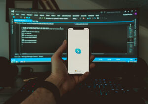 Person holding a smartphone displaying Skype in front of a computer