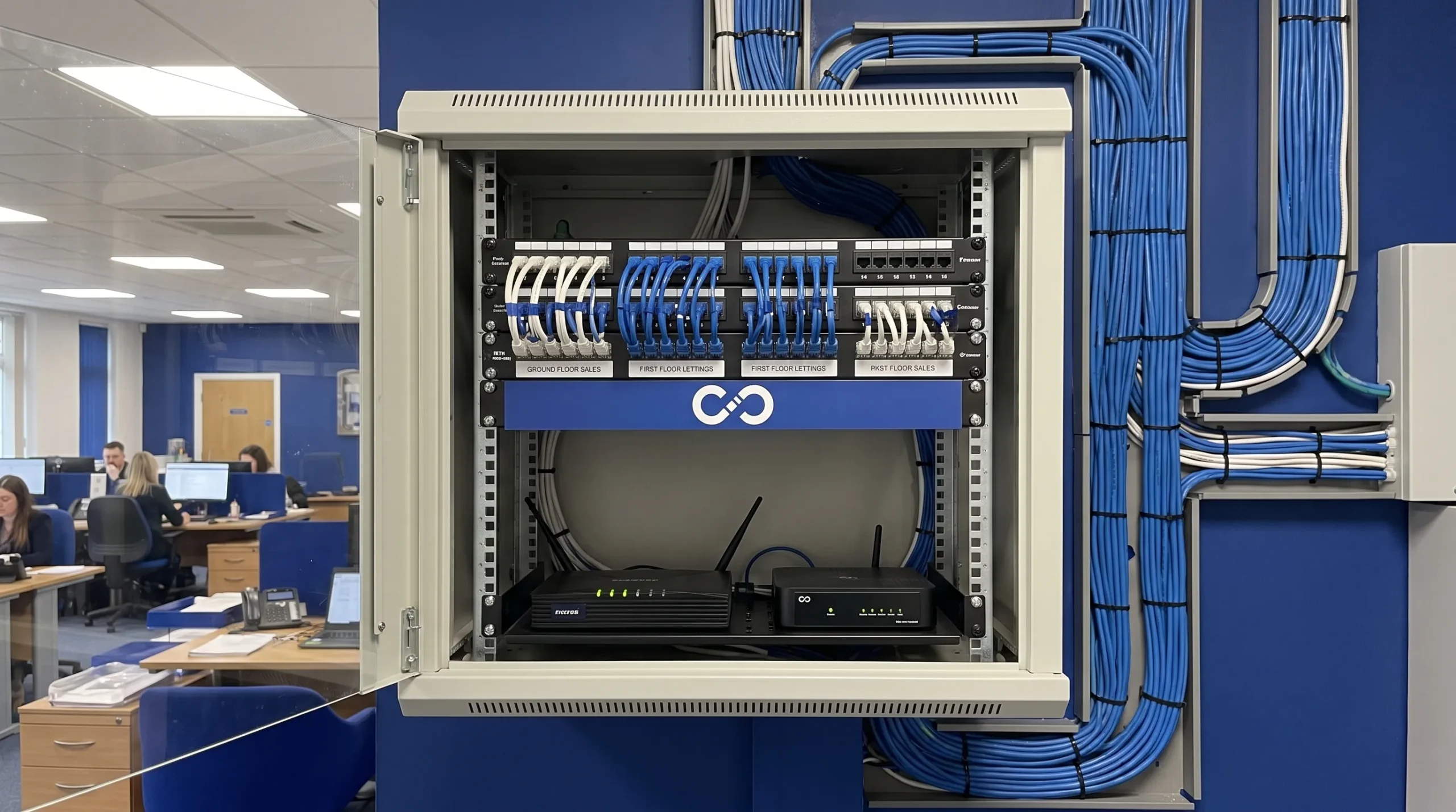A labelled network cabinet with a backup internet router supports IT support for estate agents across branch offices.
